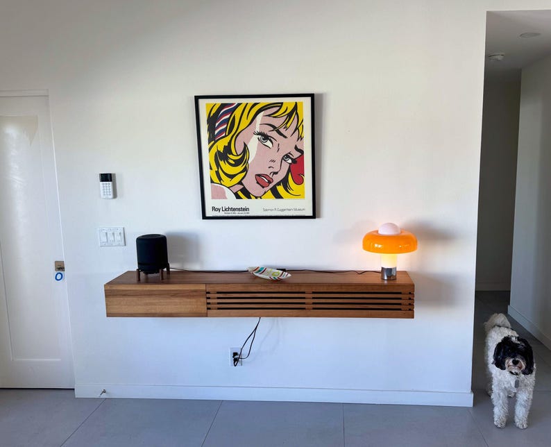 May include: A floating shelf with a black speaker, a small plate, and an orange and white lamp. A framed print of a comic book style woman's face by Roy Lichtenstein hangs above the shelf. A white dog is standing in the doorway.