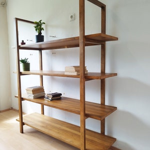 May include: A wooden bookshelf with four shelves and a rectangular frame. The shelves are made of solid wood and are stained a warm brown color. The bookshelf is against a white wall and has small plants and books on the shelves.