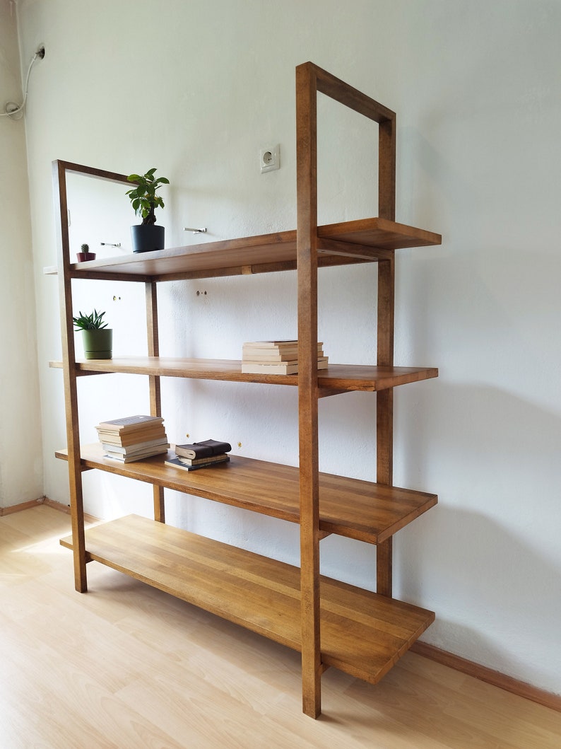 May include: A wooden bookshelf with four shelves and a rectangular frame. The shelves are made of solid wood and are stained a warm brown color. The bookshelf is against a white wall and has small plants and books on the shelves.