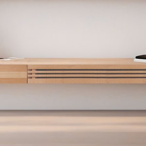 May include: A light wood floating shelf with a drawer on the left and three black and brown horizontal lines on the right. The shelf is mounted on a white wall and has a small wooden sculpture and a black object on top.