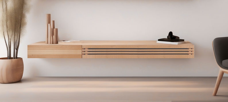 May include: A light wood floating shelf with a drawer on the left and three black and brown horizontal lines on the right. The shelf is mounted on a white wall and has a small wooden sculpture and a black object on top.