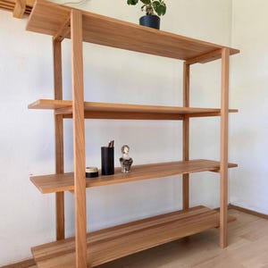 May include: A light brown wooden bookshelf with four shelves. The shelves are supported by vertical posts. Two small potted plants and a few decorative items are displayed on the shelves. The bookshelf is made of natural wood.