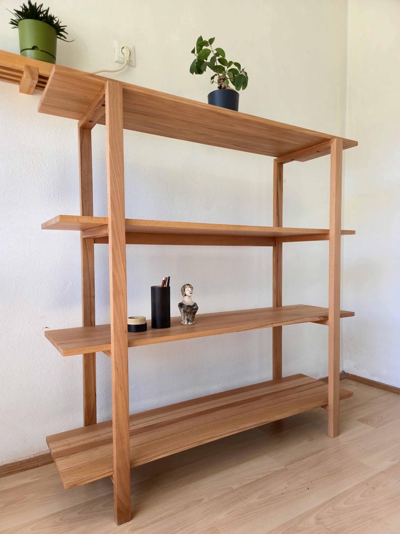 May include: A light brown wooden bookshelf with four shelves. The shelves are supported by vertical posts. Two small potted plants and a few decorative items are displayed on the shelves. The bookshelf is made of natural wood.