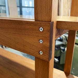 May include: Close-up of a wooden bookshelf with visible joinery. The shelves and supports are made of light brown wood, with screws securing the joints. The design features a simple, sturdy construction, showcasing the natural wood grain.