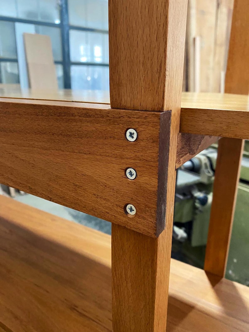 May include: Close-up of a wooden bookshelf with visible joinery. The shelves and supports are made of light brown wood, with screws securing the joints. The design features a simple, sturdy construction, showcasing the natural wood grain.