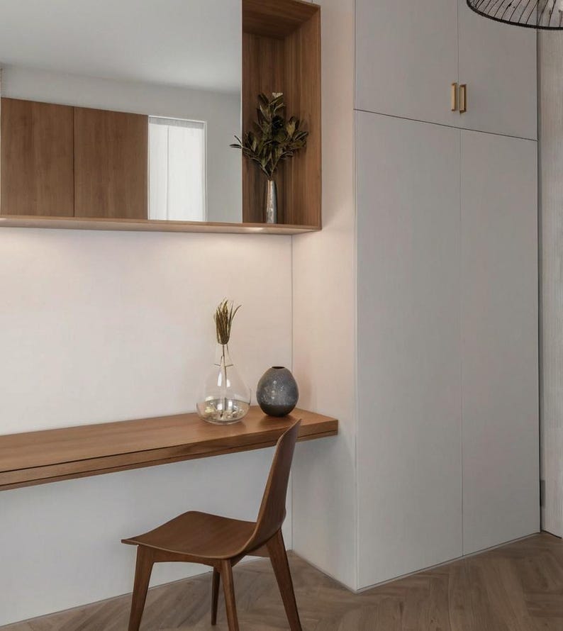 May include: A wooden desk with a white wall and a chair. The desk has a shelf above it with a vase of dried flowers. There is a small vase with dried flowers on the desk. The wall has a white cabinet with a gold door handle labeled "10".