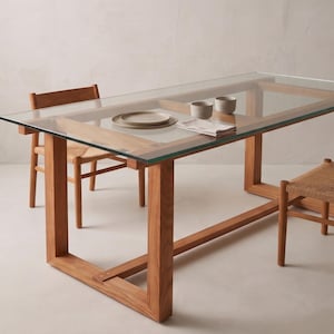 Puede incluir: Una mesa de comedor rectangular con una tapa de vidrio transparente y un marco de madera. El marco es de madera marrón claro. Se ven dos sillas con asientos tejidos. Un plato, dos cuencos y una servilleta están sobre la mesa.