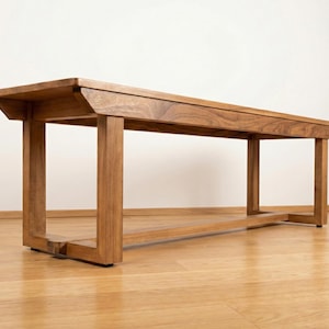 May include: A wooden bench with a rectangular top and four sturdy legs. The bench is made of light brown wood and has a simple, modern design. The bench is approximately 60 inches long.