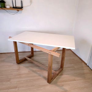 K&ouml;nnte beinhalten: Ein moderner Schreibtisch mit wei&szlig;er Tischplatte und einem Rahmen aus Naturholz. Der Schreibtisch hat eine rechteckige Oberseite und abgewinkelte Beine, wodurch ein minimalistisches Design entsteht. Der Schreibtisch ist ca. 127 cm lang und 76 cm breit.