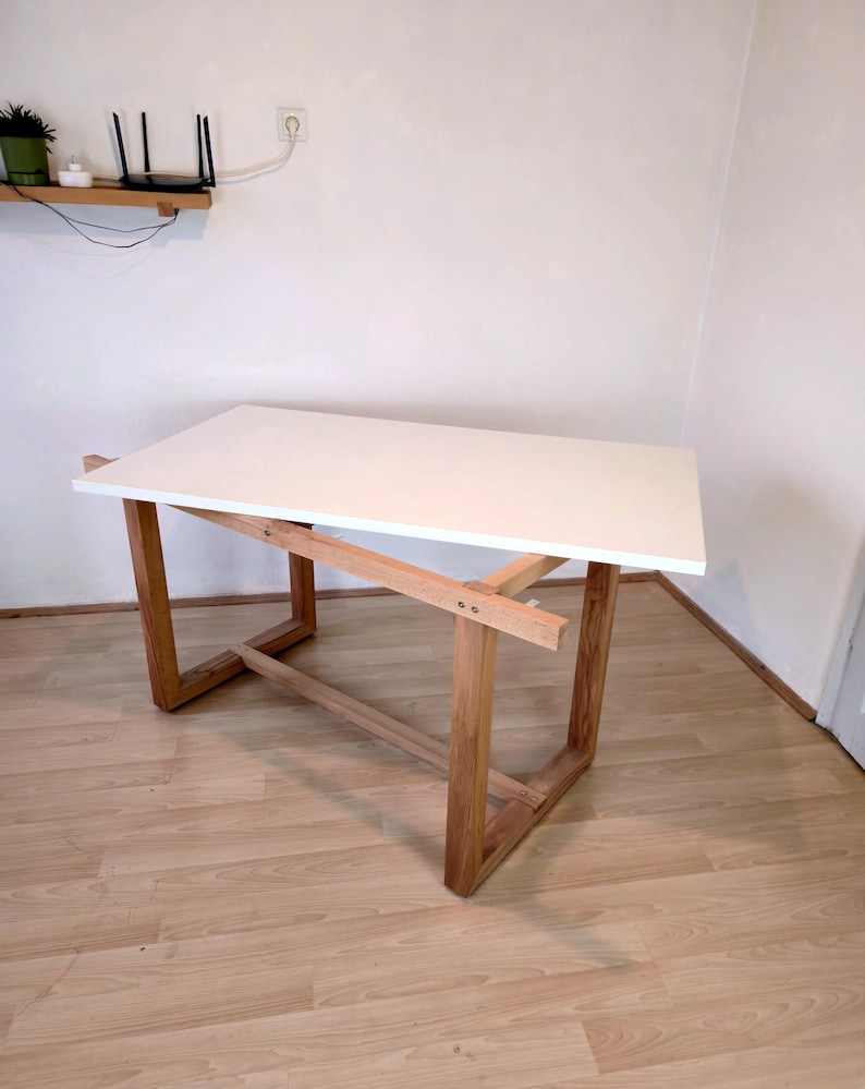 K&ouml;nnte beinhalten: Ein moderner Schreibtisch mit wei&szlig;er Tischplatte und einem Rahmen aus Naturholz. Der Schreibtisch hat eine rechteckige Oberseite und abgewinkelte Beine, wodurch ein minimalistisches Design entsteht. Der Schreibtisch ist ca. 127 cm lang und 76 cm breit.