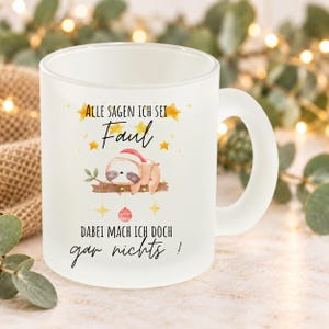 Könnte beinhalten: Ein mattierter Glasbecher mit einer Aquarellillustration eines Faultiers mit roter Weihnachtsmütze auf einem Ast. Der Text lautet: Alle sagen ich sei Faul, dabei mach ich doch gar nichts! Verziert mit gelben Sternen und Weihnachtskugeln.