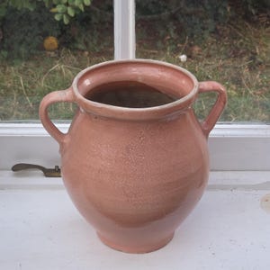 May include: A terracotta-coloured ceramic pot with a rounded body, a wide circular opening, and two symmetrical side handles. The surface features a smooth, glossy glaze with subtle variations in tone and a slightly textured appearance.