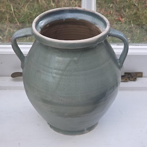May include: A ceramic vessel with a rounded body and two side handles, featuring a smooth, glossy sage green glaze. The pot has a wide, circular opening with a thick rim and a tapered base, showcasing subtle horizontal ridges along the exterior surface.