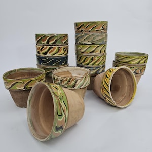 May include: Terracotta plant pots with a decorative, swirled rim design in green, brown, and yellow. Some pots are stacked, others are arranged individually, showcasing the varied patterns. The pots are suitable for indoor or outdoor use.