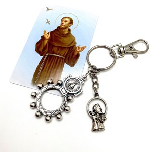 May include: Silver keyring with a Saint Francis of Assisi charm, a rosary ring, and a small cross charm. The keyring is attached to a silver key ring. The charm features a depiction of Saint Francis holding a bird in his hand. The rosary ring has a silver loop and 12 silver beads. The cross charm has a silver loop and a small silver cross with a depiction of the Virgin Mary.