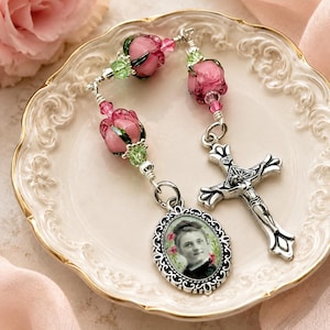 May include: A rosary featuring pink rose-shaped beads, small green crystal accents, and silver-toned metal components. The piece includes a silver crucifix and an oval-shaped pendant with a black and white portrait of a person framed in ornate silver.