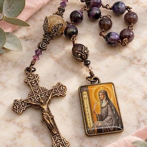 May include: A rosary featuring purple and brown stone beads, small purple crystals, and a large yellow bead in a decorative metal cap. It includes an ornate bronze-toned crucifix and a rectangular pendant depicting St. Hildegard.