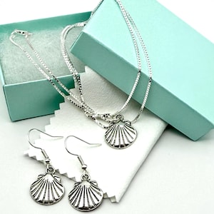 May include: A silver-toned necklace and matching earrings set. The necklace features a delicate chain with a seashell pendant. The earrings also have seashell charms. The jewelry is displayed in a light blue gift box.