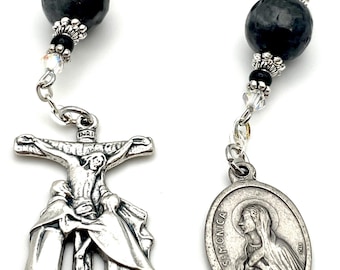 Op de afbeelding: Een zilveren rozenkrans met een kruisbeeld met een afbeelding van Jezus aan het kruis met twee figuren aan weerszijden. De rozenkrans heeft zwarte en heldere kralen en een zilveren ovale hanger met de tekst "St. Monica".