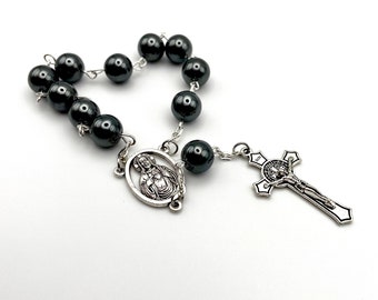 May include: A black rosary with a silver crucifix and a silver charm featuring a religious figure. The rosary beads are made of black hematite.