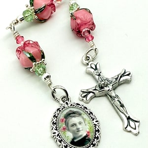 May include: A rosary featuring pink rose-shaped beads, green accents, and a silver-toned cross pendant. The rosary includes an oval pendant with a portrait and a decorative border. The beads are strung on silver wire.