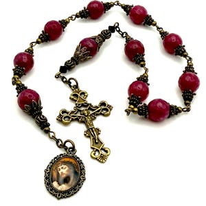 May include: A burgundy and gold rosary with a cross and a pendant featuring an image of Jesus. The rosary beads are made of faceted stone.