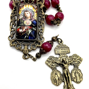 Può includere: Un rosario color borgogna e oro con un'immagine incorniciata della Vergine Maria e un crocifisso con l'iscrizione "JESUS NAZARENUS REX JUDAEORUM".