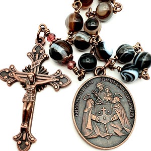 Puede incluir: Un rosario con cuentas de ágata marrón y negra y detalles metálicos color cobre. El rosario incluye un crucifijo con la figura de Jesús y una medalla con figuras religiosas y texto.