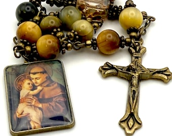 May include: A rosary with olive green, brown, and yellow beads. The rosary features a metal crucifix and a rectangular pendant with an image of a saint holding a child. The beads are strung on a metal chain.