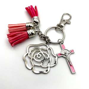 Può includere: Un portachiavi argentato con un ciondolo a forma di rosa rosa, un pon pon rosa e bianco e una croce argentata con intarsi in smalto rosa e una rappresentazione di Gesù.