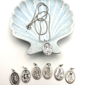 Könnte beinhalten: Ein Satz von acht silbernen ovalen Anhängern mit religiösen Motiven. Die Anhänger sind in einer Reihe auf einer weißen Oberfläche angeordnet. Die Anhänger sind alle gleich groß und gleich geformt, aber jeder hat ein anderes Bild. Die Bilder zeigen eine Frau mit einer Krone, eine Frau mit einem Baby, eine Frau mit einem Kreuz, eine Frau mit einem Herzen, eine Frau mit einer Taube, eine Frau mit einem Stern, eine Frau mit einer Sonne und eine Frau mit einem Mond.