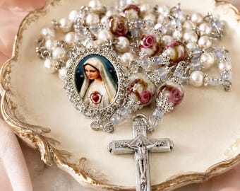 May include: A religious rosary featuring white pearl beads, clear crystal spacers, and floral patterned glass beads. The centerpiece is an oval medal depicting the Virgin Mary with a sacred heart, and the crucifix is silver-toned metal.