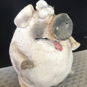 Könnte beinhalten: Weiße Keramik-Sparbüchse mit schwarzer Nase, roter Zunge und bemalten Augen. Die Sparbüchse ist leicht abgebrochen und hat einen abgenutzten Look.