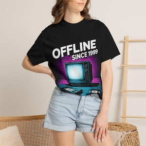 Könnte beinhalten: Schwarzes T-Shirt mit der Aufschrift "OFFLINE SINCE 1999" über einer Retro-Fernsehgrafik. Die Grafik enthält einen lila und blauen Backsteinmauer-Hintergrund und zwei VHS-Kassetten. Das T-Shirt hat einen Rundhalsausschnitt.