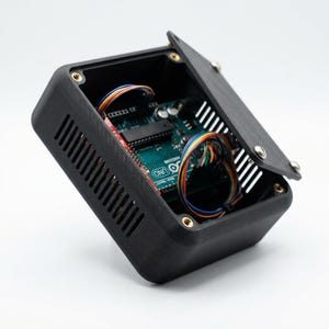 May include: A black rectangular enclosure with a hinged lid, featuring side ventilation slots and brass threaded inserts. Inside, a green Arduino Uno circuit board is visible, connected with multi-colored wiring bundles throughout the interior.