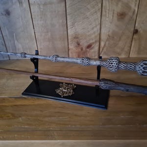 May include: Two detailed wands, one light gray and the other brown, are displayed on a black stand. The light gray wand has a textured design, while the brown wand has a textured handle. A gold emblem is on the stand.