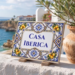 May include: A rectangular ceramic tile sign featuring the text CASA IBERICA in blue lettering. The border is decorated with intricate blue and yellow floral patterns and illustrations of yellow lemons on a white background.