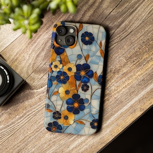 Può includere: Una custodia per telefono con un motivo floreale blu e giallo. La custodia ha uno sfondo bianco e un design a mosaico. La custodia per telefono ha un ritaglio per una fotocamera a tre obiettivi.