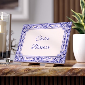May include: A square ceramic tile with a blue and white floral border and the words "Casa Blanca" in blue script. The tile is propped up on a wooden stand, and is on a marble surface.