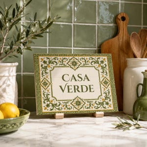 Peut inclure: Une plaque rectangulaire en céramique portant l'inscription CASA VERDE en lettres vert foncé. La plaque est ornée d'une bordure complexe composée de vignes vertes, de fleurs jaunes et de motifs géométriques sur un fond crème.
