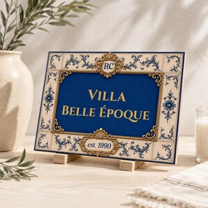May include: A rectangular sign featuring a deep blue centre with the gold text Villa Belle Époque and est. 1990. The border is cream-coloured with blue floral patterns and ornate gold filigree accents, including a top crest with the letters RC.