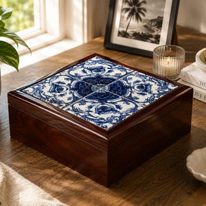 May include: A square, wooden jewelry box with a dark brown finish. The lid is adorned with four square tiles featuring a blue and white floral pattern. The box sits on a wooden surface, with a plant and framed picture in the background.