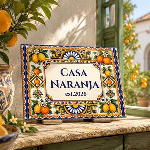 May include: A rectangular ceramic tile sign featuring a cream-coloured centre with the dark blue text "CASA NARANJA est.2026". The border is decorated with a colourful pattern of yellow lemons, orange fruits, green leaves, and blue floral motifs.