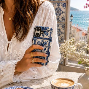 May include: A blue and white patterned phone case is held in front of a white blouse. The phone case has a floral design. A matching blue and white patterned teacup and saucer are on the table, along with a pastry on a plate.