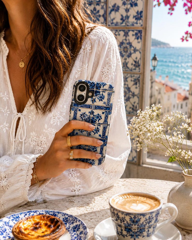 May include: A blue and white patterned phone case is held in front of a white blouse. The phone case has a floral design. A matching blue and white patterned teacup and saucer are on the table, along with a pastry on a plate.
