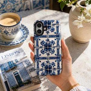 May include: A blue and white patterned phone case, reminiscent of traditional Portuguese tiles, is held in a hand. The case features a floral design and is set against a backdrop of a coffee cup, saucer, and a book titled "Lisbon Soul of a City."