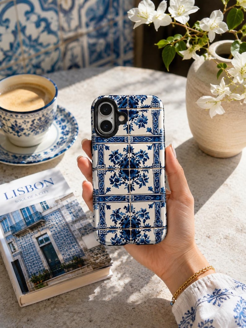 May include: A blue and white patterned phone case, reminiscent of traditional Portuguese tiles, is held in a hand. The case features a floral design and is set against a backdrop of a coffee cup, saucer, and a book titled "Lisbon Soul of a City."