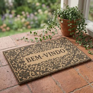 Puede incluir: Felpudo rectangular con fondo beige y borde floral y de volutas marrón oscuro. El texto "BEM-VINDO" está centrado en la alfombra. Una planta en maceta se encuentra cerca sobre una superficie de ladrillo.