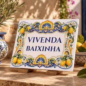May include: A rectangular ceramic tile sign featuring the text VIVENDA BAIXINHA in dark blue lettering. The border is decorated with yellow lemons, green leaves, white flowers, and ornate blue and yellow scrollwork patterns on a white background.