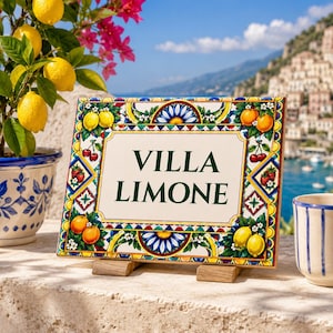 以下が含まれることがあります： VILLA LIMONEという濃い緑色の文字が書かれた長方形のセラミック製看板。縁取りには、黄色いレモン、赤いサクランボ、イチゴ、青い花柄の手描きのモチーフが白地にあしらわれています。